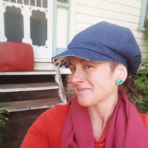 May include: A denim newsboy cap with a patterned lining. The cap is worn with a red shirt and a burgundy scarf. A red suitcase sits on the steps of a house. The person is wearing turquoise earrings.