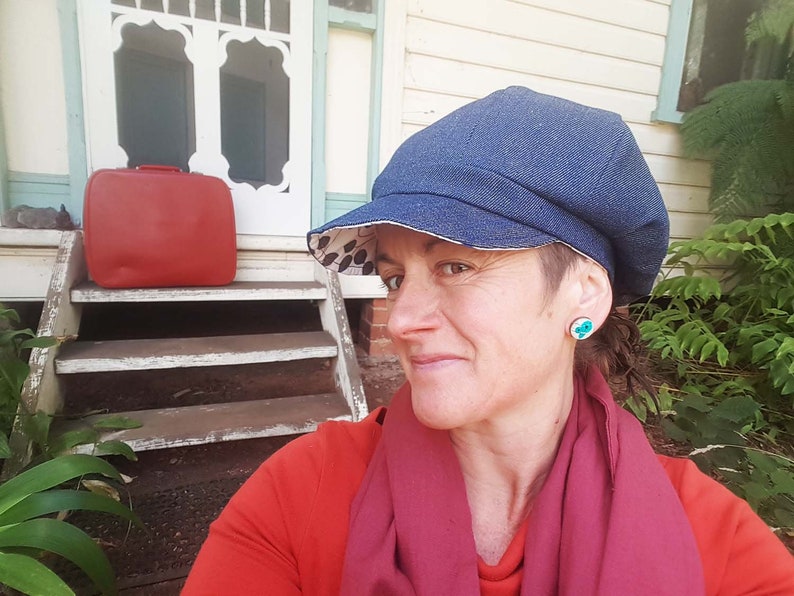 May include: A denim newsboy cap with a patterned lining. The cap is worn with a red shirt and a burgundy scarf. A red suitcase sits on the steps of a house. The person is wearing turquoise earrings.