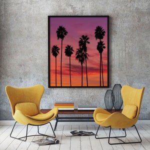 May include: A framed print of a sunset with palm trees silhouetted against the sky. The sky is a gradient of pink, orange, and purple. The palm trees are black and are arranged in a row.
