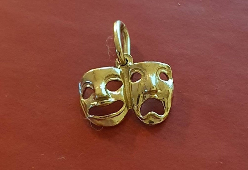 May include: A gold-colored pendant charm featuring two theatrical masks, one smiling and one frowning.