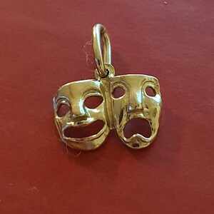 May include: A gold-colored pendant charm featuring two theater masks, one smiling and one frowning. The masks are connected at the top and have a loop for attaching to a chain.