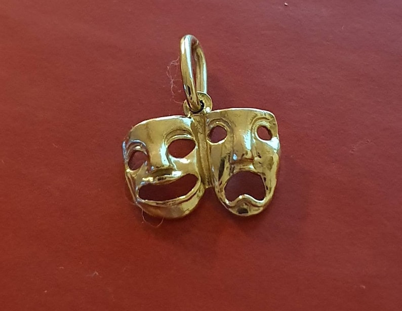 May include: A gold-colored pendant charm featuring two theater masks, one smiling and one frowning. The masks are connected at the top and have a loop for attaching to a chain.