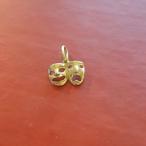May include: A gold-colored pendant charm featuring two theatrical masks, one smiling and one frowning. The masks are joined together and have a loop at the top for attaching to a chain.