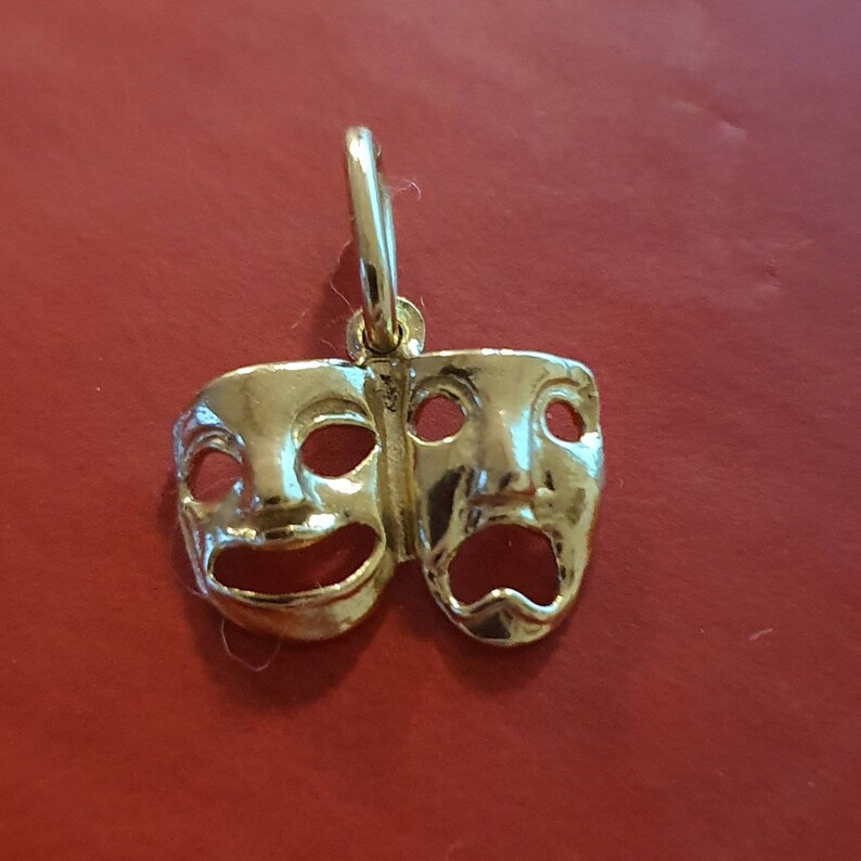 May include: A gold-colored pendant featuring two theatrical masks, one smiling and one frowning. The masks are joined together at the top and have a loop for hanging.