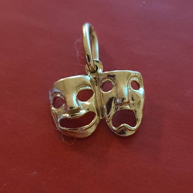 May include: A gold-colored pendant charm featuring two theatrical masks, one smiling and one frowning. The masks are joined together at the top and have a loop at the top for attaching to a chain.