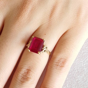 May include: A gold ring featuring a large, emerald-cut ruby gemstone. The ring has a delicate, ornate design.