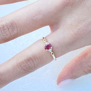 May include: A gold ring featuring a large, round, red gemstone, flanked by two smaller, clear gemstones. The ring has a delicate, twisted band design. The ring is shown on a finger.