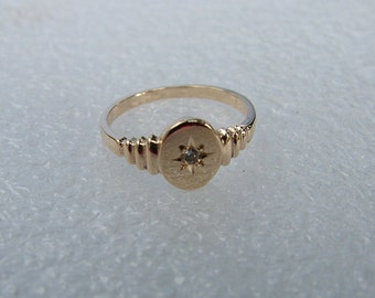 Könnte beinhalten: Ein goldfarbener Ring mit einem Sternmuster auf einer ovalen Fläche. Der Ring hat ein schmales Band und dekorative Rillen auf beiden Seiten der Fläche. Ein kleiner, klarer Stein ist in der Mitte des Sterns gesetzt.