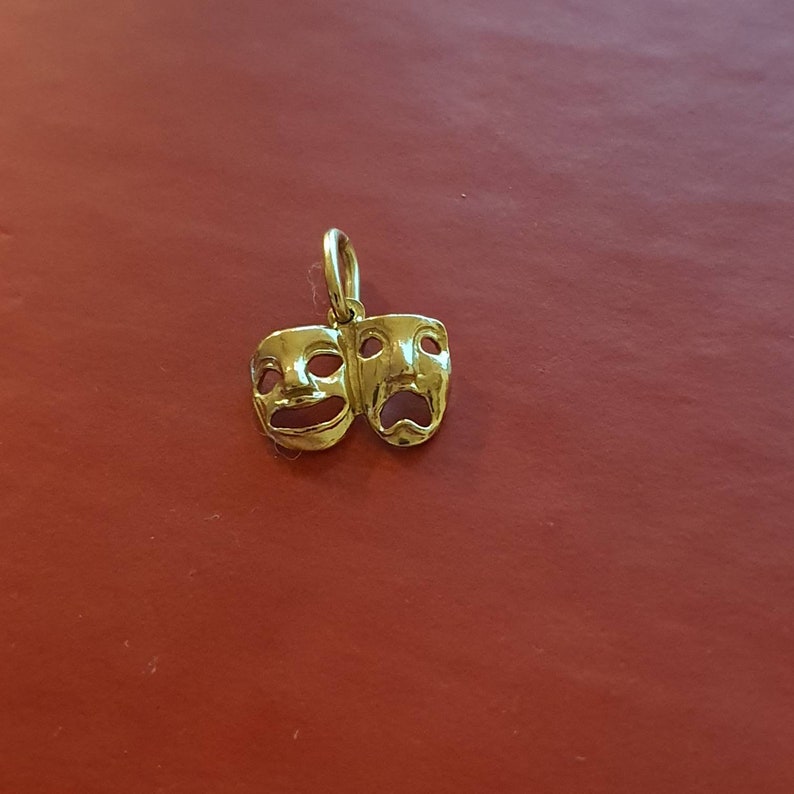 May include: A gold-colored charm featuring two theatrical masks, one smiling and one frowning. The masks are joined together at the top and have a loop at the top for hanging.
