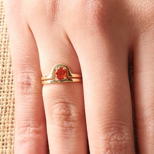 May include: A gold ring with a red gemstone set in a curved band. The ring is worn on a person's finger.