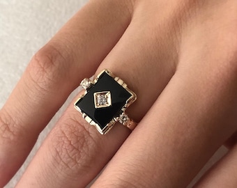 May include: A gold ring featuring a black square stone with smaller diamonds surrounding it.