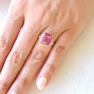 May include: A gold ring featuring a large, pink, emerald-cut gemstone. The ring is set with smaller diamonds around the gemstone.
