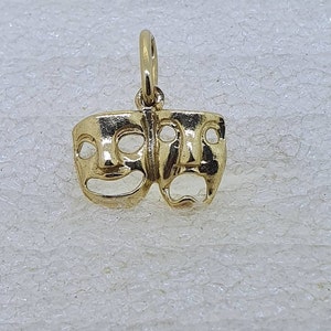 May include: A gold-colored charm featuring two theatrical masks, one smiling and one frowning. The masks are connected at the top and have a loop for attaching to a necklace or bracelet.