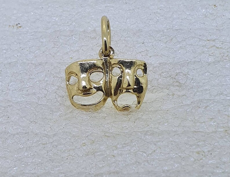 May include: A gold-colored charm featuring two theatrical masks, one smiling and one frowning. The masks are connected at the top and have a loop for attaching to a necklace or bracelet.