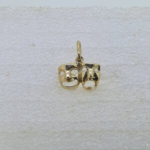 May include: A gold-colored pendant charm featuring two stylized faces, one smiling and one frowning. The charm is a classic theatrical symbol representing comedy and tragedy.