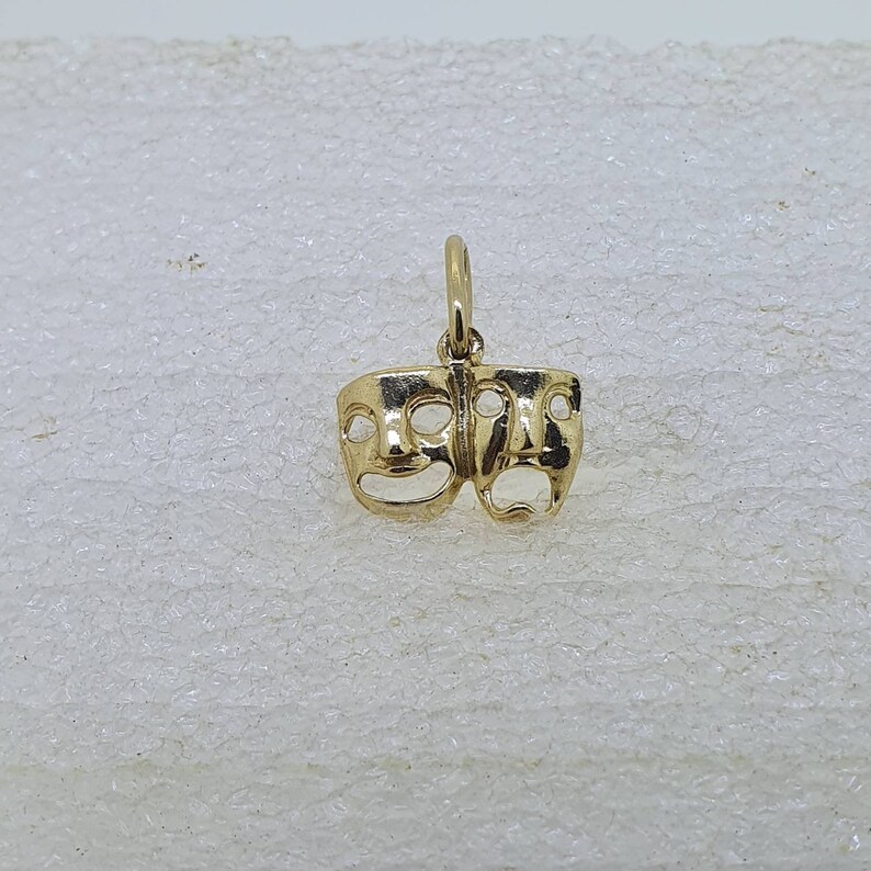May include: A gold-colored pendant charm featuring two stylized faces, one smiling and one frowning. The charm is a classic theatrical symbol representing comedy and tragedy.
