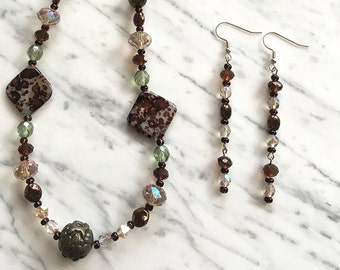 Könnte beinhalten: Eine braune und grüne Perlenkette mit zwei passenden Ohrringen. Die Kette hat zwei quadratische, braune und weiße Perlen mit Muster und eine runde, braune Perle. Die Ohrringe bestehen aus braunen und klaren Perlen.