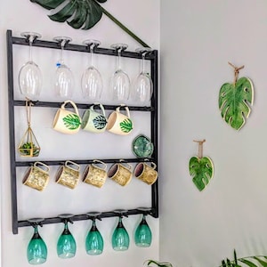 May include: A black metal wall-mounted rack with hooks for hanging wine glasses and mugs. The rack has two shelves and is decorated with green and white ceramic mugs featuring tropical leaf designs. There are also green ceramic leaf-shaped wall decorations.