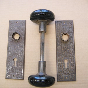 May include: Antique door hardware set featuring a black glass doorknob and two ornate metal backplates. The doorknob has a dark, glossy finish, and the backplates have keyholes and decorative floral patterns. A vintage home decor item.