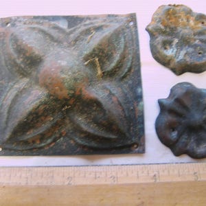 May include: Aged metal decorative hardware pieces. Includes a square plate with a floral design and two smaller, matching floral-shaped pieces. A wooden ruler is included for scale.