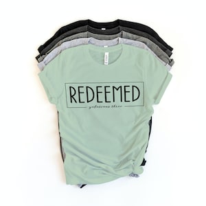 May include: A light green t-shirt with the word "REDEEMED" in black text and the words "galatians three" in smaller black text below. The shirt is folded and stacked with other t-shirts in gray and black.
