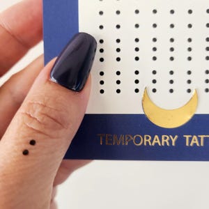 Può includere: Un tatuaggio temporaneo con due punti neri, che rappresentano un punto e virgola, sul dito di una persona. Il tatuaggio è su una carta bianca con punti neri e una luna crescente dorata.