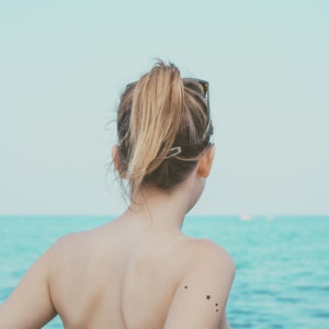 May include: A woman with long blonde hair pulled back in a ponytail stands facing away from the camera. She is wearing a black swimsuit and has a small star tattoo on her shoulder. The woman is standing on a beach with the ocean in the background.