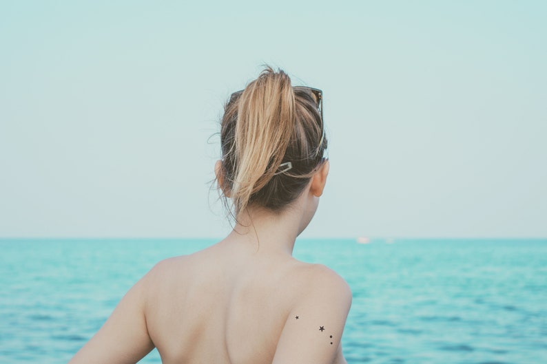 May include: A woman with long blonde hair pulled back in a ponytail stands facing away from the camera. She is wearing a black swimsuit and has a small star tattoo on her shoulder. The woman is standing on a beach with the ocean in the background.