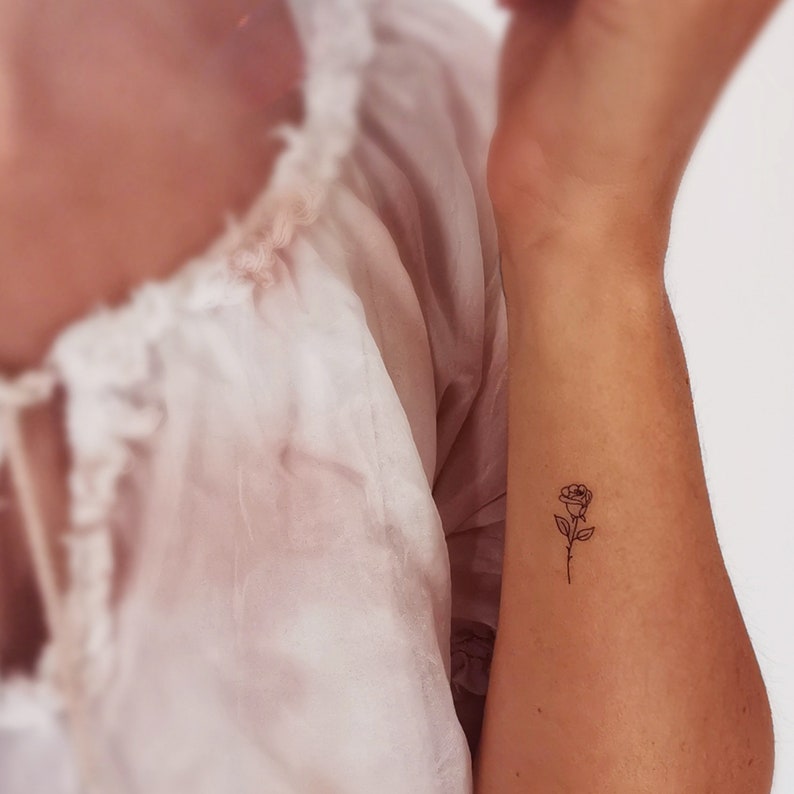 May include: A simple line drawing of a rose tattoo on a person's wrist.