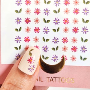 May include: A sheet of nail tattoos with a crescent moon design and a variety of small floral designs in pink, purple, and green. The sheet is labeled "Nail Tattoos".
