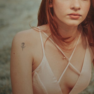 May include: A woman with long red hair wearing a white tank top with a delicate rose tattoo on her shoulder.