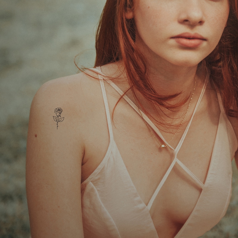 May include: A woman with long red hair wearing a white tank top with a delicate rose tattoo on her shoulder.