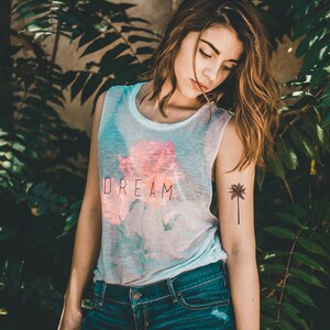 May include: A woman wearing a white vest top with a pink rose and the word "DREAM" printed on it. She is also wearing blue denim shorts and has a palm tree tattoo on her arm.