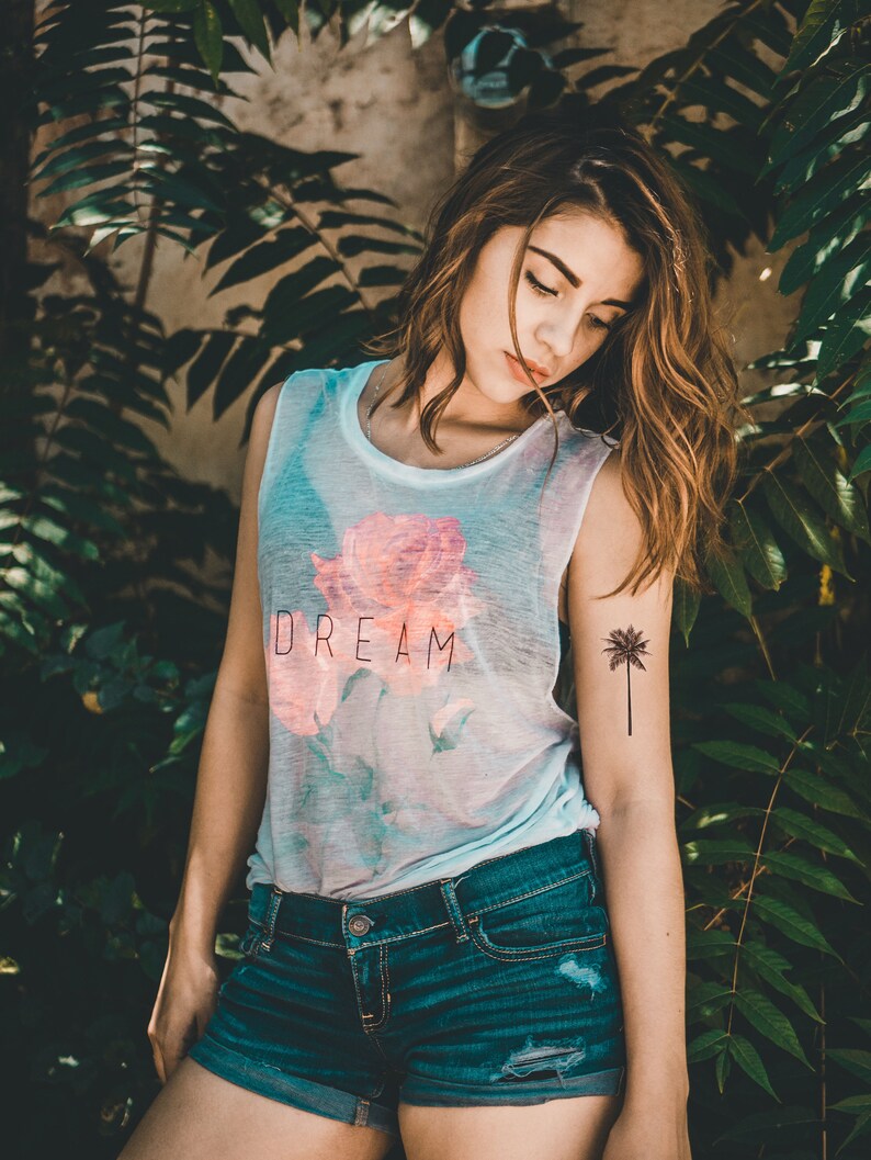 May include: A woman wearing a white vest top with a pink rose and the word "DREAM" printed on it. She is also wearing blue denim shorts and has a palm tree tattoo on her arm.