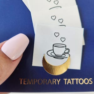May include: A set of temporary tattoos featuring a black and white illustration of a cup of tea with hearts above it and a gold crescent moon below. The text "TEMPORARY TATTOOS" is printed on the blue background.