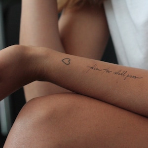 Könnte beinhalten: Nahaufnahme eines Arms mit zwei Tätowierungen. Eines ist ein kleines Herz, das andere der kursive Satz "this too shall pass". Der Hautton ist hell, und die Tätowierungen sind in schwarzer Tinte.
