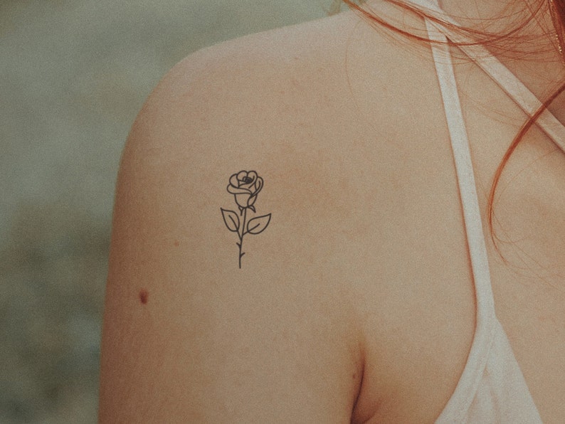 May include: A simple line drawing of a rose with a stem and leaves, tattooed on someone's shoulder.