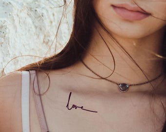May include: A close-up of a woman's collarbone with a simple "love" tattoo in cursive script. She is wearing a delicate silver necklace with a small circular pendant.