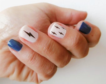 Pode incluir: Uma mão com esmalte de unha branco com desenhos de raios pretos nas unhas. Duas das unhas estão pintadas de uma cor azul escura.