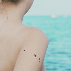 May include: A person with a small star tattoo on their shoulder, with two smaller dots below it. The person is facing away from the camera and is standing in front of a body of water.