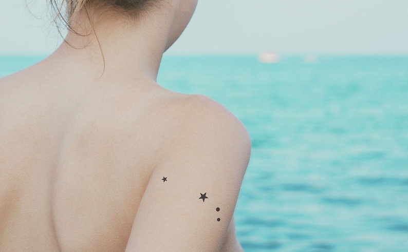 May include: A person with a small star tattoo on their shoulder, with two smaller dots below it. The person is facing away from the camera and is standing in front of a body of water.