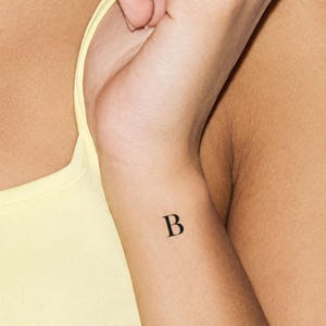 May include: A small, black, serif-style letter B temporary tattoo applied to the skin of a wrist. The letter is rendered in a classic, clean typeface with distinct serifs, positioned centrally on the inner forearm area near the wrist joint.