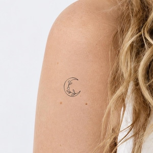 Pode incluir: Uma pequena tatuagem temporária preta apresentando o contorno de uma lua crescente com um delicado design de ramo floral no interior. A tatuagem está aplicada na pele, exibindo um estilo de arte de linha fina com dois pequenos botões.