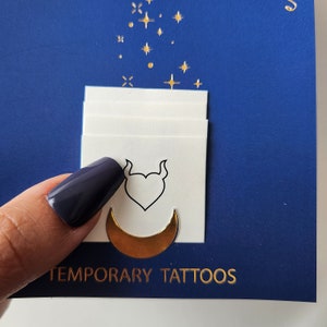 May include: A pack of temporary tattoos with a gold crescent moon and a heart with horns design. The tattoos are on a white background and the text "TEMPORARY TATTOOS" is printed in gold on a blue background.
