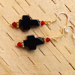 May include: A pair of black cross-shaped earrings with gold-coloured hooks. The earrings feature red and gold-coloured beads. The earrings are displayed on a light brown wooden surface.