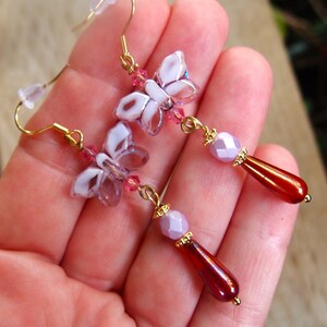 May include: Elegant butterfly earrings with gold-coloured hooks. Each earring features a butterfly made of white and lavender petals, pink beads, a lavender bead, and a teardrop-shaped, dark red bead.