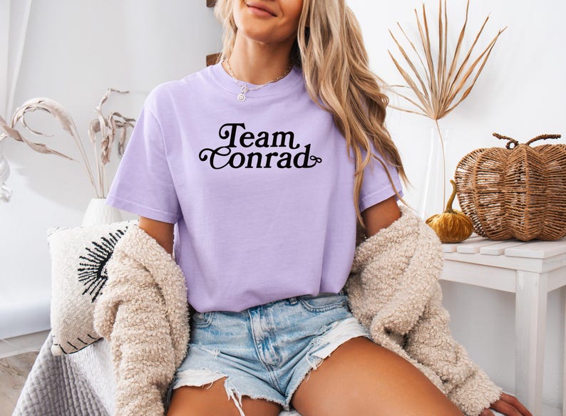 May include: A light purple t-shirt with the text "Team Conrad" in a black, cursive font. The shirt is short-sleeved and appears to be made of a soft material. The model is wearing denim shorts and a cream-colored cardigan.