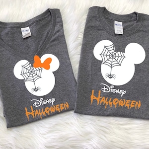 May include: Two gray t-shirts with a white Mickey Mouse head design. One shirt has a spider web and spider on the head and the text "Disney Halloween" in orange. The other shirt has a spider web, spider, and a bow on the head and the text "Disney Halloween" in orange.