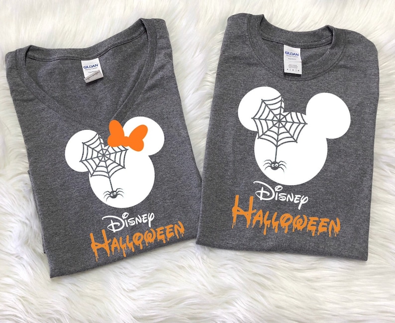 May include: Two gray t-shirts with a white Mickey Mouse head design. One shirt has a spider web and spider on the head and the text "Disney Halloween" in orange. The other shirt has a spider web, spider, and a bow on the head and the text "Disney Halloween" in orange.