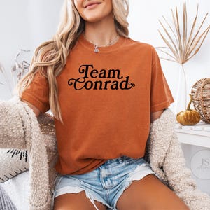 May include: An orange t-shirt with the text "Team Conrad" in a black script font. The shirt is worn with denim shorts and a cream-colored cardigan. A decorative pumpkin and woven basket are in the background.